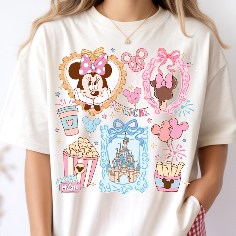 Puede incluir: Camiseta color crema con ilustraciones de dibujos animados coloridos de Minnie Mouse, un castillo, palomitas de ma&iacute;z y otros elementos de parques tem&aacute;ticos. La palabra "MAGICAL" tambi&eacute;n est&aacute; impresa en la camiseta.