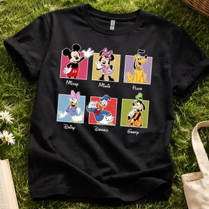 May include: Black t-shirt featuring six Disney characters: Mickey, Minnie, Pluto, Daisy, Donald, and Goofy. Each character is in a colorful square with their name below. The shirt is on a green grassy surface.