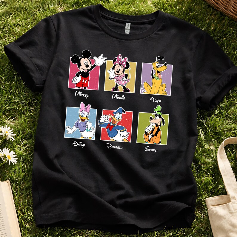 May include: Black t-shirt featuring six Disney characters: Mickey, Minnie, Pluto, Daisy, Donald, and Goofy. Each character is in a colorful square with their name below. The shirt is on a green grassy surface.
