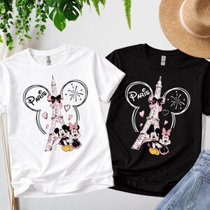 K&ouml;nnte beinhalten: Zwei T-Shirts, eines wei&szlig; und eines schwarz, mit einem Design mit dem Eiffelturm, Mickey und Minnie Mouse und dem Wort "Paris". Das Design umfasst rosa Herzen, Schleifen und Feuerwerk.
