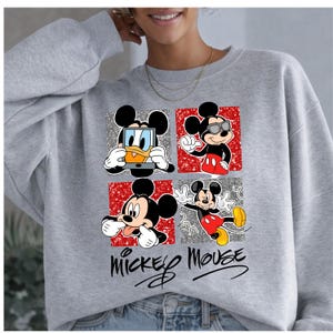 Op de afbeelding: Een grijze sweatshirt met een vierpaneelsontwerp met Mickey Mouse in verschillende poses. Elk paneel heeft een unieke achtergrond, waaronder zilveren glitter en rode patronen. De woorden "Mickey Mouse" staan onderaan in cursief.