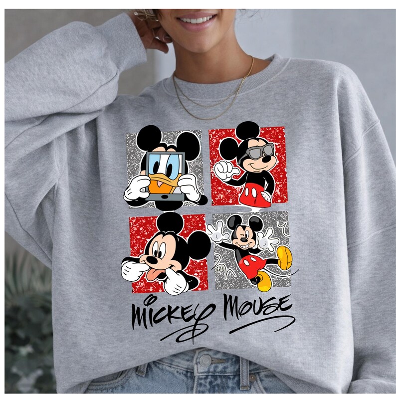 Op de afbeelding: Een grijze sweatshirt met een vierpaneelsontwerp met Mickey Mouse in verschillende poses. Elk paneel heeft een unieke achtergrond, waaronder zilveren glitter en rode patronen. De woorden "Mickey Mouse" staan onderaan in cursief.