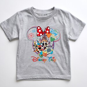 Puede incluir: Una camiseta gris con un gr&aacute;fico colorido de personajes de Disney en un coche frente a un castillo. El gr&aacute;fico incluye un lazo rojo, fuegos artificiales y las palabras "Best Day" y "Disney Trip".
