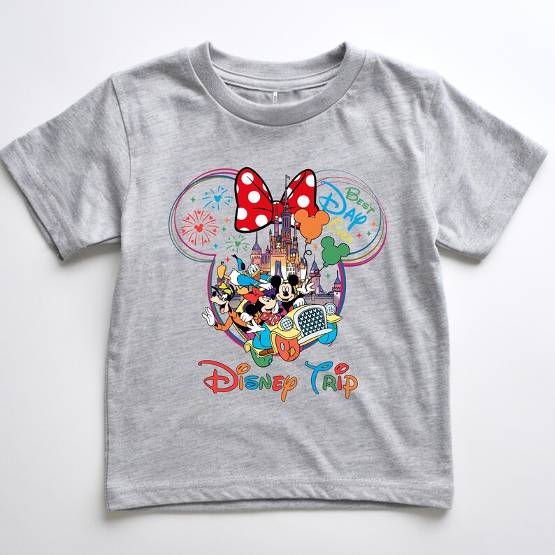 Puede incluir: Una camiseta gris con un gr&aacute;fico colorido de personajes de Disney en un coche frente a un castillo. El gr&aacute;fico incluye un lazo rojo, fuegos artificiales y las palabras "Best Day" y "Disney Trip".