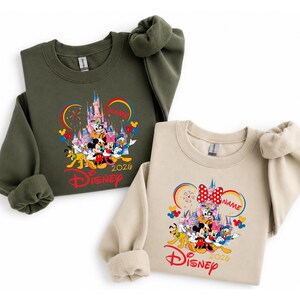 Op de afbeelding: Twee sweatshirts, &eacute;&eacute;n olijfgroen en &eacute;&eacute;n beige, met een kleurrijke Disney-afbeelding. De afbeelding toont Disney-personages, een kasteel, vuurwerk en het woord "Disney" in het rood. Het jaar "2026" is ook zichtbaar.