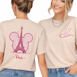 May include: Beige t-shirts with pink graphics. One shirt features the Eiffel Tower inside a Mickey Mouse head outline with the word "Paris". The other shirt has a castle graphic. Both shirts have short sleeves.