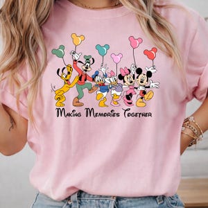 May include: Pink t-shirt featuring a graphic of Disney characters including Mickey Mouse, Minnie Mouse, Donald Duck, Daisy Duck, Goofy, and Pluto. The characters are holding Mickey Mouse-shaped balloons. The text "Making Memories Together" is below the characters.