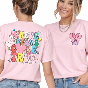 May include: Pale pink t-shirts with a colourful graphic design. One shirt displays the text "WHERE DREAMS COME TRUE" with Mickey Mouse-themed elements. The other shirt has a Minnie Mouse bow and castle design.