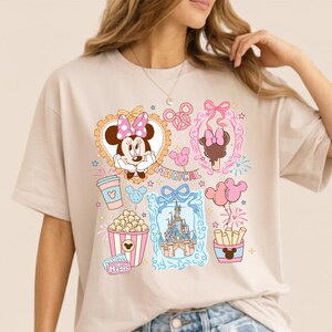 Puede incluir: Camiseta beige con un dise&ntilde;o gr&aacute;fico colorido con Minnie Mouse, un castillo, palomitas de ma&iacute;z y otros elementos de parques tem&aacute;ticos. Tambi&eacute;n se muestra la palabra "MAGICAL". La camiseta tiene mangas cortas y un corte holgado.