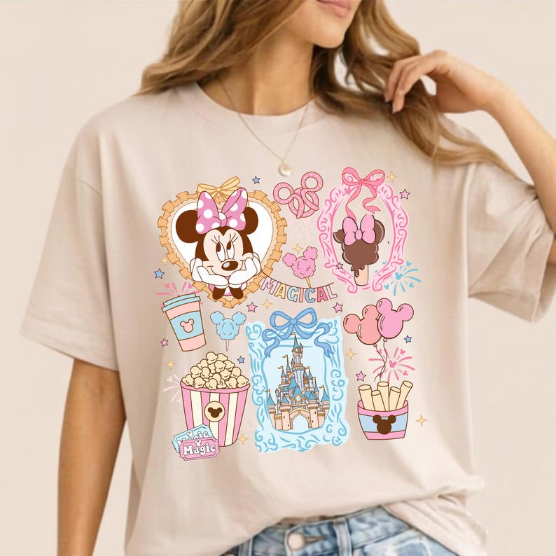 Puede incluir: Camiseta beige con un dise&ntilde;o gr&aacute;fico colorido con Minnie Mouse, un castillo, palomitas de ma&iacute;z y otros elementos de parques tem&aacute;ticos. Tambi&eacute;n se muestra la palabra "MAGICAL". La camiseta tiene mangas cortas y un corte holgado.