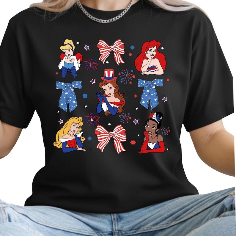 May include: Black t-shirt with cartoon illustrations of princesses, patriotic bows, fireworks, and stars. The princesses are in red, white, and blue, celebrating a festive theme.