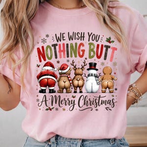 May include: A light pink t-shirt featuring a graphic of Santa Claus, a bear, a reindeer, a snowman, and a gingerbread person facing away. The text reads WE WISH YOU NOTHING BUTT A MERRY CHRISTMAS, surrounded by small snowflakes and holly accents.