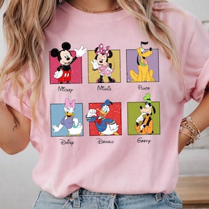 May include: A light pink t-shirt featuring six Disney cartoon characters in colorful squares. Mickey Mouse, Minnie Mouse, Pluto, Daisy Duck, Donald Duck, and Goofy are displayed with their names below. The shirt has rolled-up sleeves.