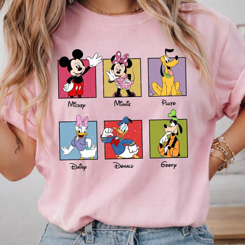 May include: A light pink t-shirt featuring six Disney cartoon characters in colorful squares. Mickey Mouse, Minnie Mouse, Pluto, Daisy Duck, Donald Duck, and Goofy are displayed with their names below. The shirt has rolled-up sleeves.