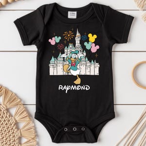 Może przedstawiać: Czarny body niemowlęce z kolorową grafiką Kaczora Donalda przed zamkiem, fajerwerkami i balonami. Imię "Raymond" jest wydrukowane na dole na biało. Body ma kr&oacute;tkie rękawy i trzy zatrzaski na dole.