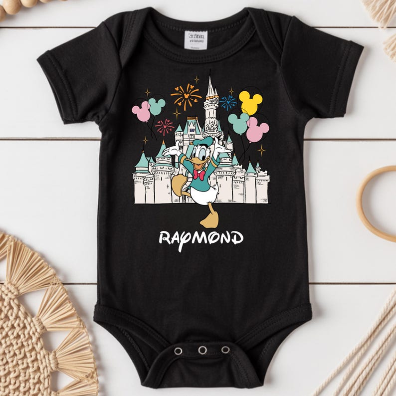Może przedstawiać: Czarny body niemowlęce z kolorową grafiką Kaczora Donalda przed zamkiem, fajerwerkami i balonami. Imię "Raymond" jest wydrukowane na dole na biało. Body ma kr&oacute;tkie rękawy i trzy zatrzaski na dole.