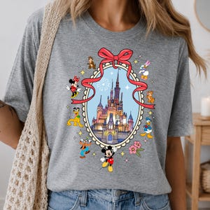 May include: A gray t-shirt featuring a colorful oval design with a castle, fireworks, and various cartoon characters. The design includes Mickey Mouse, Minnie Mouse, Donald Duck, and other characters, with a red bow at the top.