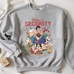 May include: A grey sweatshirt with a graphic of cartoon characters and the text "Princess Security Club" in red. The sweatshirt has a crew neck and long sleeves. The design features various cartoon characters.