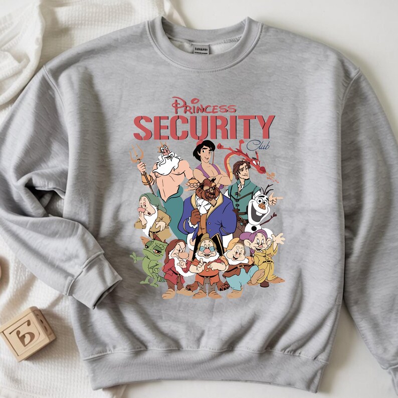 May include: A grey sweatshirt with a graphic of cartoon characters and the text "Princess Security Club" in red. The sweatshirt has a crew neck and long sleeves. The design features various cartoon characters.