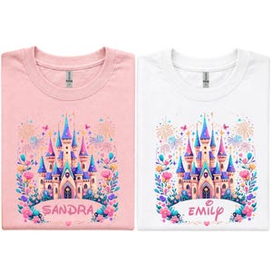 K&ouml;nnte beinhalten: Zwei T-Shirts, eines rosa und eines wei&szlig;en, jeweils mit einem farbenfrohen Schlossdesign. Das Schloss hat blaue, rosa und lila T&uuml;rme, umgeben von Blumen und Feuerwerk. Das rosa Shirt sagt "SANDRA" und das wei&szlig;e Shirt sagt "EMILY".