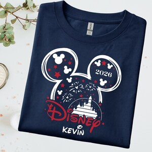 May include: Navy blue t-shirt with a white and red Mickey Mouse head design. The design includes red stars, fireworks, and the text "2026" and "Disney Kevin". The shirt is made of a soft material.