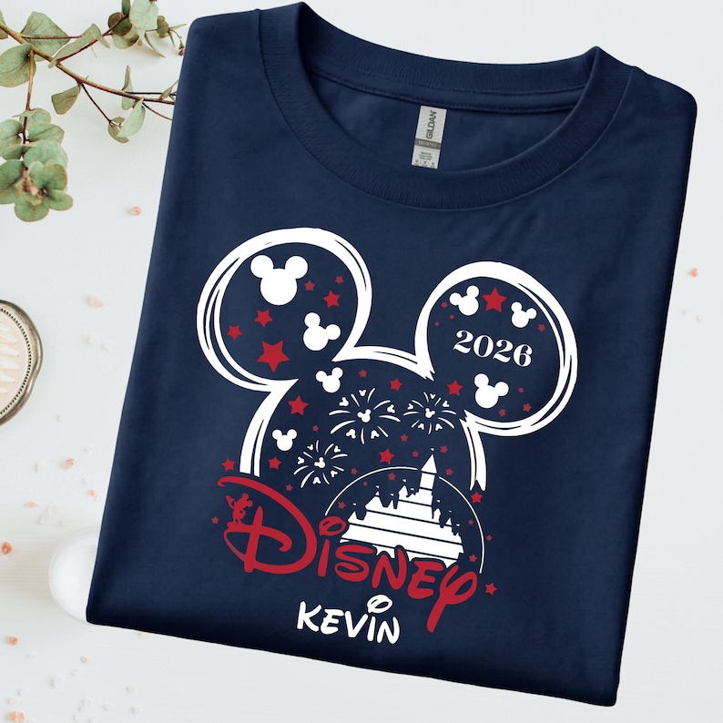 May include: Navy blue t-shirt with a white and red Mickey Mouse head design. The design includes red stars, fireworks, and the text "2026" and "Disney Kevin". The shirt is made of a soft material.