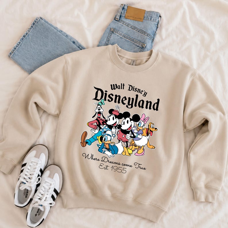 Pu&ograve; includere: Felpa girocollo beige con una grafica colorata dei personaggi Disney, tra cui Topolino e Minnie, Paperino e Pippo. Il testo "Walt Disney Disneyland" e "Where Dreams come True Est. 1955" sono stampati sopra e sotto la grafica.