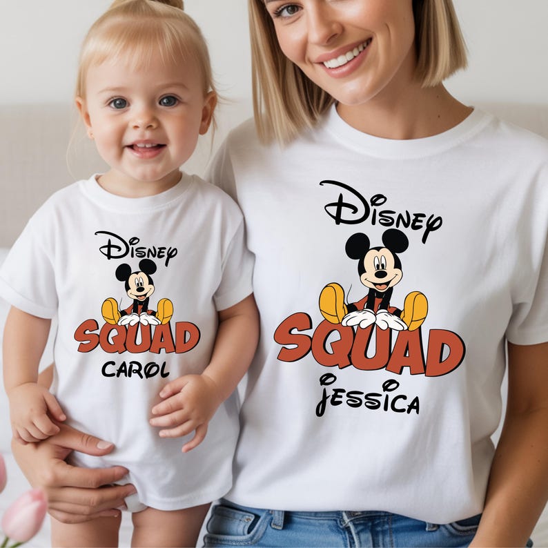 Op de afbeelding: Witte T-shirts met een Disney Mickey Mouse-ontwerp. De shirts tonen de woorden "Disney Squad" in oranje en zwart, met gepersonaliseerde namen eronder. E&eacute;n shirt heeft "Carol", de andere "Jessica".
