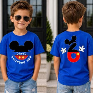 May include: A blue t-shirt with a Mickey Mouse design. The front of the shirt has Mickey's head and the words "DAVID BIRTHDAY BOY". The back of the shirt has a large number 6 with Mickey Mouse hands. The shirt is worn by a child.