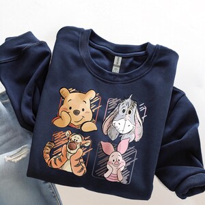 May include: Navy blue sweatshirt featuring four cartoon character portraits: Winnie the Pooh, Eeyore, Tigger, and Piglet. The illustrations are set within individual rectangular frames. The sweatshirt is folded on top of a pair of blue jeans.