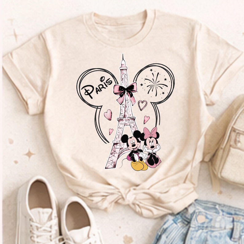 K&ouml;nnte beinhalten: Beiges T-Shirt mit einem Aufdruck des Eiffelturms, Mickey und Minnie Mouse. Das Wort "Paris" steht &uuml;ber dem Turm in einem Mickey Mouse-Ohren-Design, mit Herzen und Feuerwerk. Die Figuren sind in Schwarz, Rosa und Gelb gehalten.
