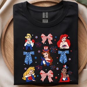 May include: Black t-shirt with a patriotic design. It features cartoon illustrations of princesses, bows, fireworks, and stars in red, white, and blue. The shirt has a crew neck and is made of a soft material.