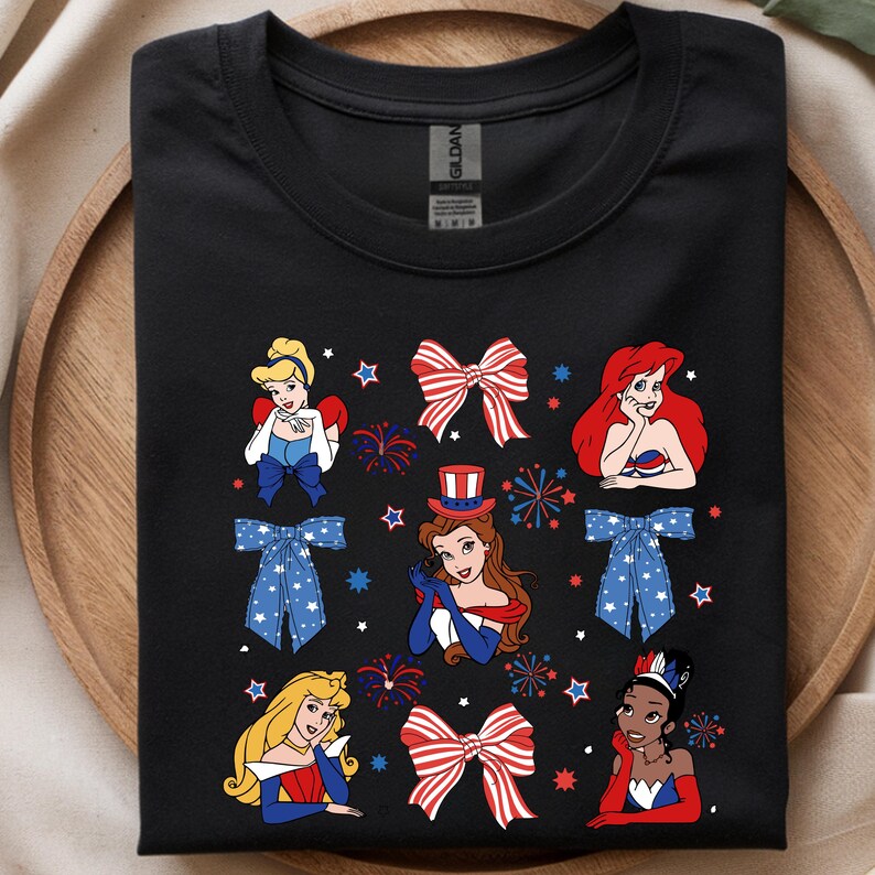 May include: Black t-shirt with a patriotic design. It features cartoon illustrations of princesses, bows, fireworks, and stars in red, white, and blue. The shirt has a crew neck and is made of a soft material.