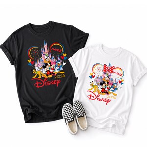Op de afbeelding: Zwarte en witte T-shirts met een Disney-ontwerp met Mickey Mouse, Minnie Mouse en andere personages. De shirts tonen het woord "Disney" in het rood, het jaar "2026" en een plek voor een naam. Een paar zwart-wit geruite instappers staat in de afbeelding.