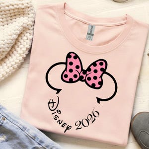 May include: A light pink t-shirt featuring a Minnie Mouse design. The design includes a black outline of Minnie's head, a pink bow with black polka dots, and the word "Disney" and the year "2026" in a script font.