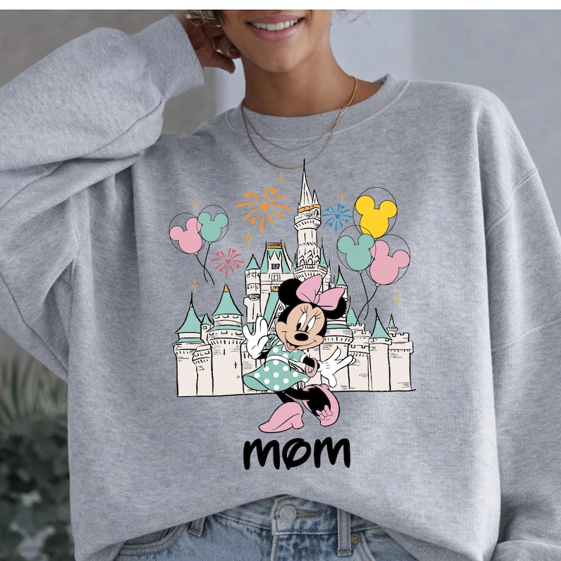 Może przedstawiać: Szara bluza z nadrukiem Myszki Minnie przed zamkiem, z balonami i fajerwerkami. Słowo "mama" jest wydrukowane poniżej. Projekt zawiera kolory r&oacute;żowy, niebieski, ż&oacute;łty i biały.