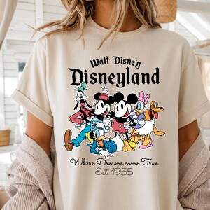 Pu&ograve; includere: T-shirt beige con la scritta "Walt Disney's Disneyland" e una grafica dei personaggi Disney, tra cui Topolino e Minnie. Il testo "Where Dreams come True Est. 1955" &egrave; sotto. La maglietta &egrave; di colore chiaro.