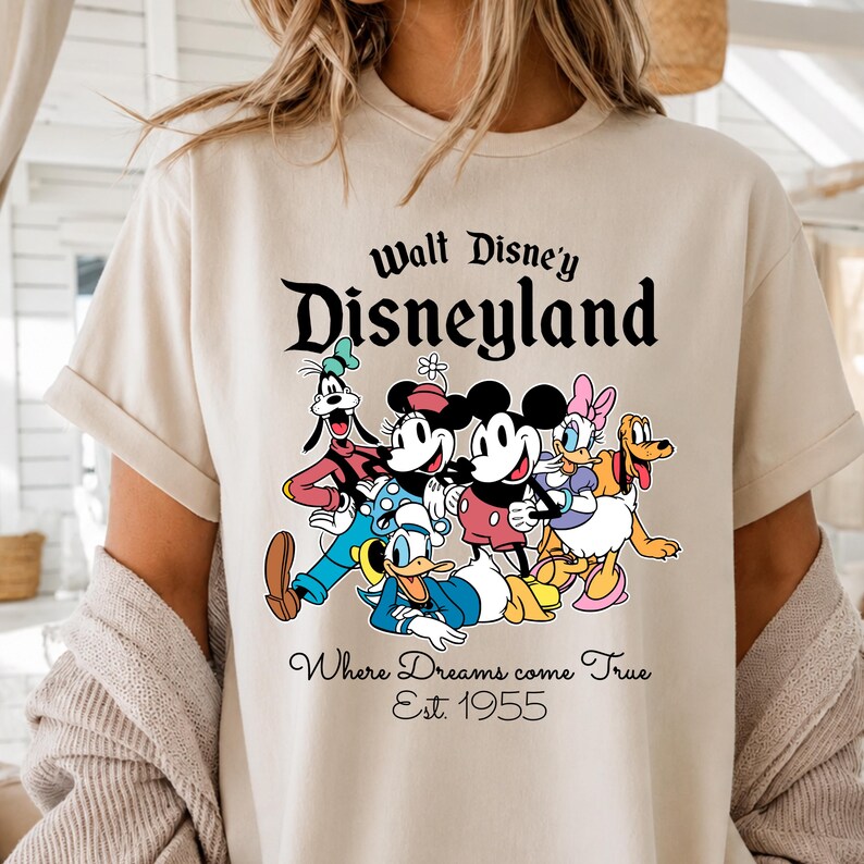 Pu&ograve; includere: T-shirt beige con la scritta "Walt Disney's Disneyland" e una grafica dei personaggi Disney, tra cui Topolino e Minnie. Il testo "Where Dreams come True Est. 1955" &egrave; sotto. La maglietta &egrave; di colore chiaro.
