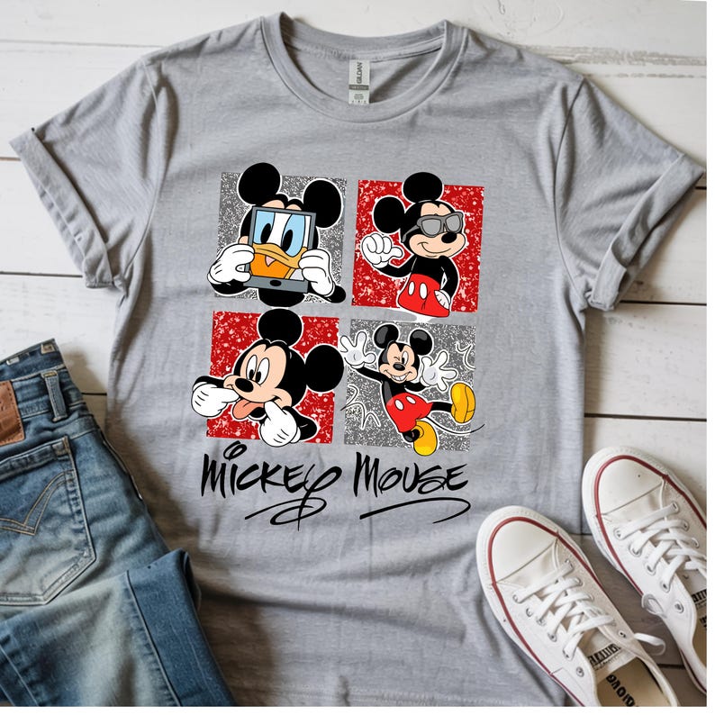 Op de afbeelding: Een grijs t-shirt met een Mickey Mouse-ontwerp. Het ontwerp bevat vier panelen met verschillende afbeeldingen van Mickey Mouse en de woorden "Mickey Mouse" in een scriptlettertype. Het shirt wordt getoond met een blauwe jeans en witte sneakers.
