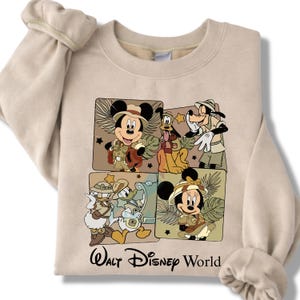Puede incluir: Sudadera beige con un gr&aacute;fico de Mickey Mouse, Pluto, Goofy, Donald Duck y Minnie Mouse con trajes de safari. Las palabras "Walt Disney World" est&aacute;n impresas en negro debajo. La sudadera tiene mangas largas.