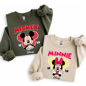 May include: Two sweatshirts, one olive green and one beige, featuring Mickey and Minnie Mouse designs. The Mickey sweatshirt has a red "MICKEY" graphic and a Mickey Mouse graphic. The Minnie sweatshirt has a pink "MINNIE" graphic and a Minnie Mouse graphic.