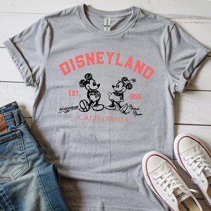 Puede incluir: Una camiseta gris con el logo de Disneyland en coral, con ilustraciones de Mickey y Minnie Mouse. La camiseta incluye el texto "EST. 1955" y "CALIFORNIA". Zapatillas blancas y vaqueros azules tambi&eacute;n son visibles.