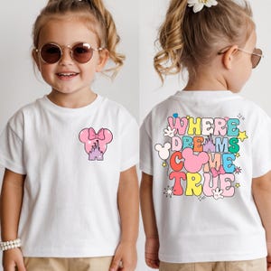 May include: White t-shirt with a pink Minnie Mouse bow and castle design on the front. The back of the shirt features the text "WHERE DREAMS COME TRUE" in colourful, playful lettering. A fun and whimsical design for children.
