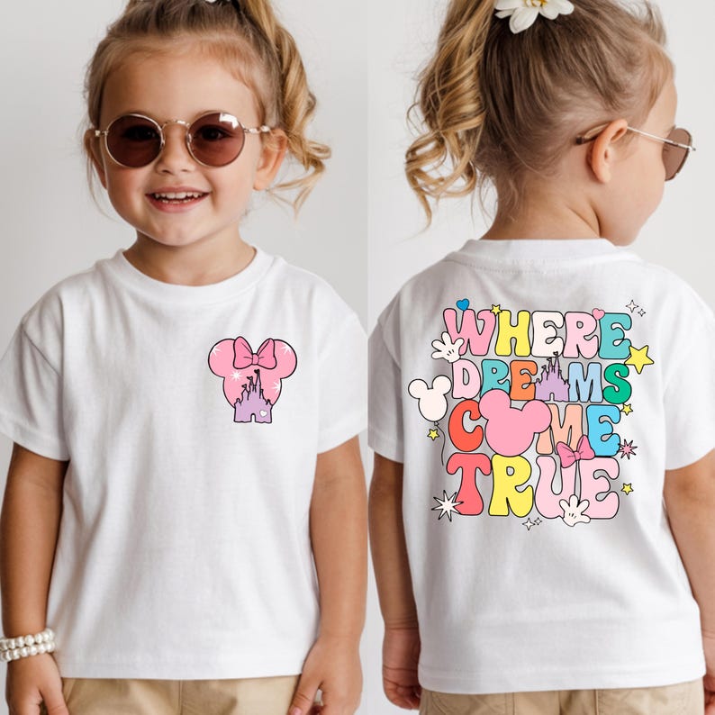 May include: White t-shirt with a pink Minnie Mouse bow and castle design on the front. The back of the shirt features the text "WHERE DREAMS COME TRUE" in colourful, playful lettering. A fun and whimsical design for children.