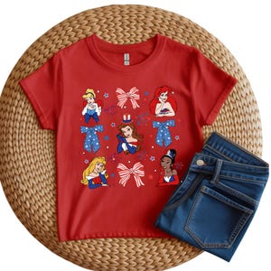 May include: A red t-shirt with cartoon princess illustrations and patriotic bows. The shirt is displayed with a pair of folded blue denim jeans. The design includes stars and fireworks, suggesting a holiday theme.