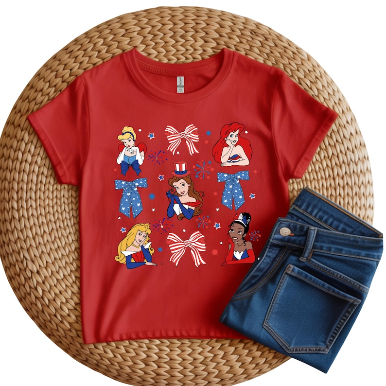 May include: A red t-shirt with cartoon princess illustrations and patriotic bows. The shirt is displayed with a pair of folded blue denim jeans. The design includes stars and fireworks, suggesting a holiday theme.
