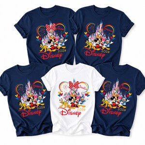 Op de afbeelding: Vijf marineblauwe en witte Disney T-shirts. Elk shirt heeft een kleurrijke afbeelding van Disney-personages, een kasteel en het woord "Disney" in het rood. De shirts hebben ook namen en het jaar 2020.