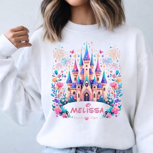 K&ouml;nnte beinhalten: Wei&szlig;es Sweatshirt mit einem farbenfrohen Schlossdesign in Rosa, Blau und Gelb. Das Design umfasst Blumen, Schmetterlinge und Feuerwerk. Der Name "MELISSA" ist darunter gedruckt, ein verspieltes Design.