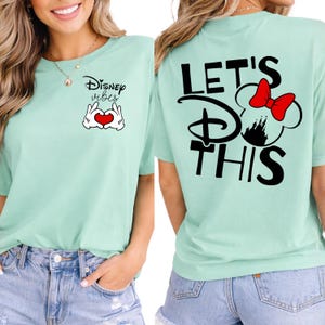 K&ouml;nnte beinhalten: Hellgr&uuml;nes T-Shirt mit dem Schriftzug "Disney vibes" und einer herzf&ouml;rmigen Handgesten-Grafik auf der Vorderseite. Auf der R&uuml;ckseite des Shirts steht der Satz "Let's Do This" mit einer Minnie-Mouse-Silhouette und einer roten Schleife.