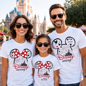 May include: White t-shirts with Disney-themed graphics. The shirts feature a Mickey Mouse head design with a red bow, fireworks, and a castle silhouette. The text includes "Disney," "Mommy," "Daddy," and a name, all in red.